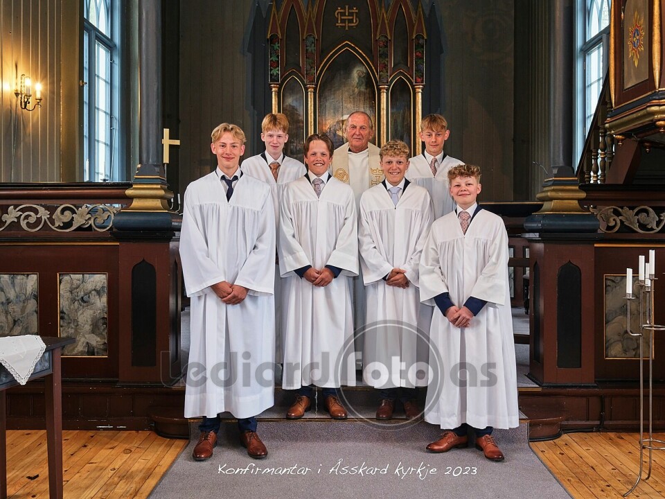 6 konfirmanter, alle gutter kledd i konfirmantkapper, oppstilt foran alteret i kirka. Bak dem står en prest.