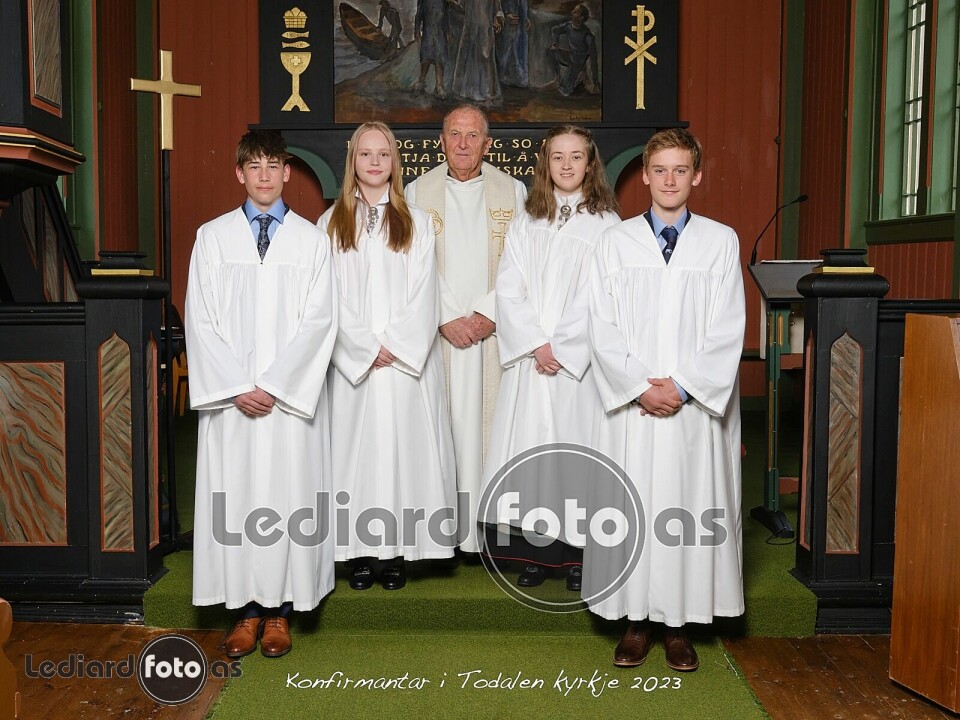 Fire konfirmanter, to jenter og to gutter, i konfirmantkapper, står oppstilt foran alteret i kirka. Bak dem står presten.