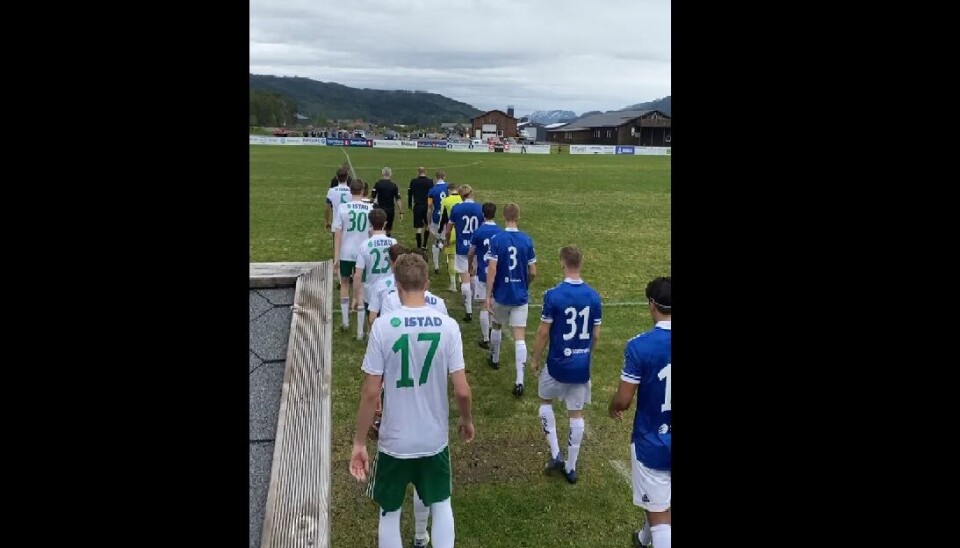 Spillere fra to lag går ut på fotballbanen. De ene med hvite og grønne drakter, det andre med blå og hvit