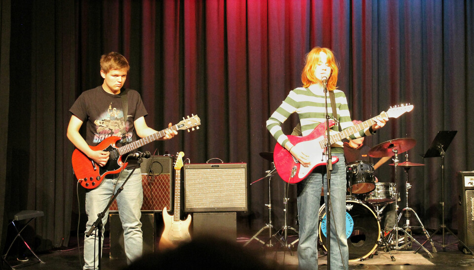To ungdommer spiller gitar og synger. I bakgrunnen spiller én på trommer.