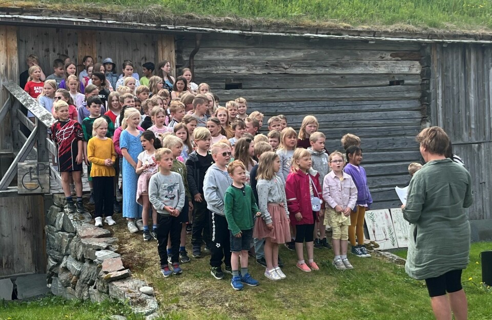 Mange barn står samlet på ei låvebru på et gammelt fjøs og synger. Læreren står foran og dirigerer.
