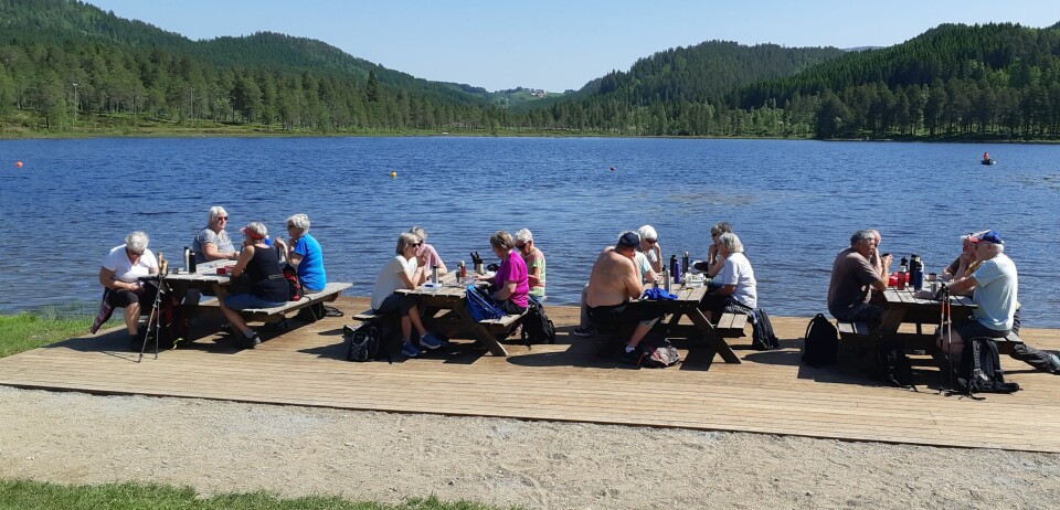 Pensjonister på tur som sitter ved flere bord på en platting ved et vann.
