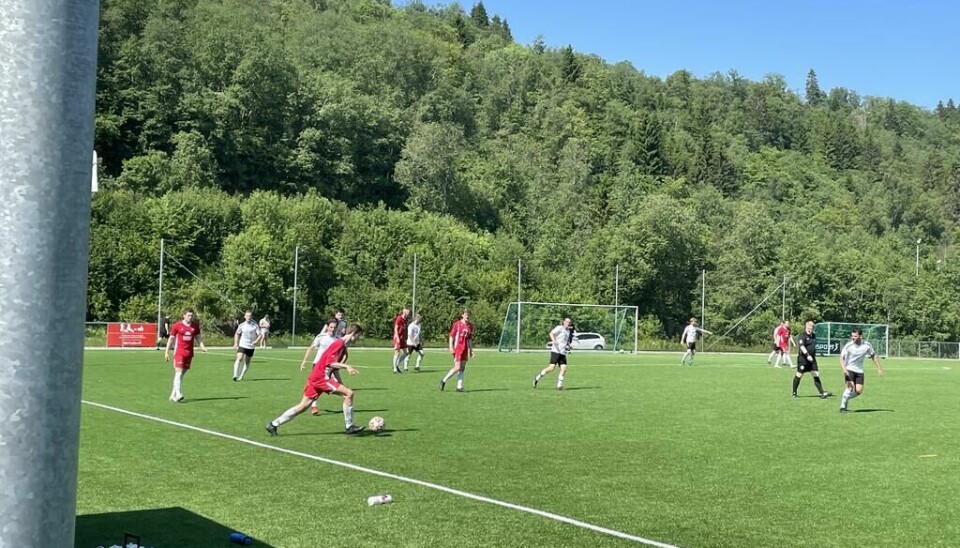 Et fotballag i røde drakter spiller mot et lag med hvite drakter på en gressbane med trær i bakgrunnen.