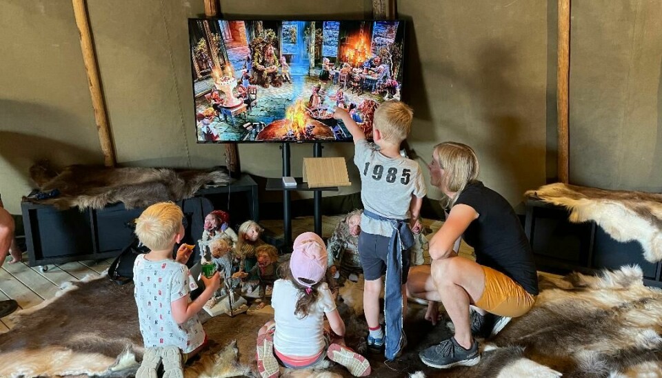 To barn og en dame sitter og ser på en skjerm med troll, mens et barn står og peker på skjermen.