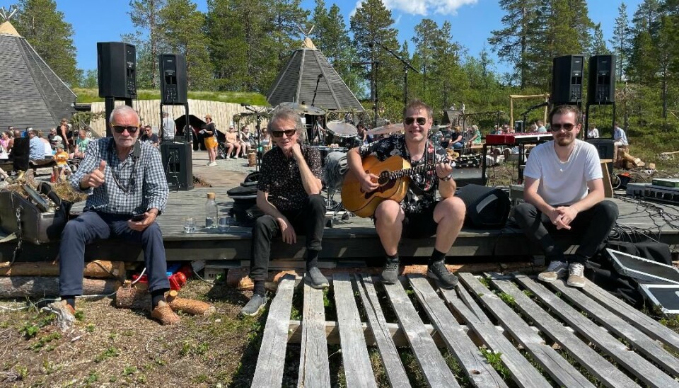 Fire menn sitter på baksiden av scenen. Den ene sitter med en gitar.