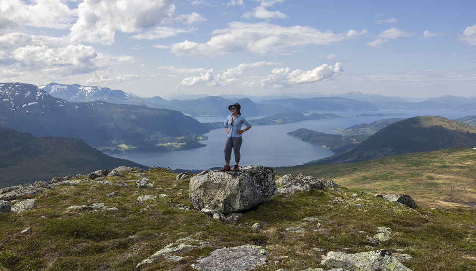 Foto fra ein tidlegare fjelltur. Ei dame med hatt som står på en stor stein på fjellet og ser seg rundt. Fjord og fjell i bakgrunnen.