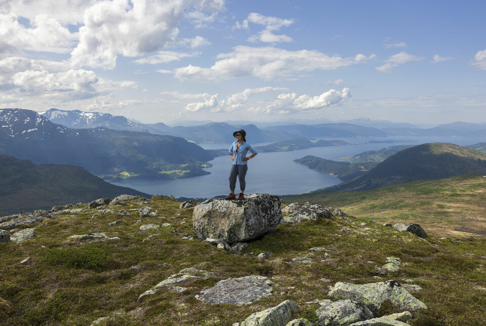 Ei dame med hatt som står på en stor stein på fjellet og ser seg rundt. Fjord og fjell i bakgrunnen.