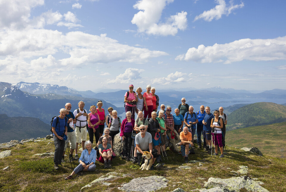 Ei gruppe med over 20 turkledde pensjonister oppstilt ved en varde på en fjelltopp. Fjord og fjell i bakgrunnen.