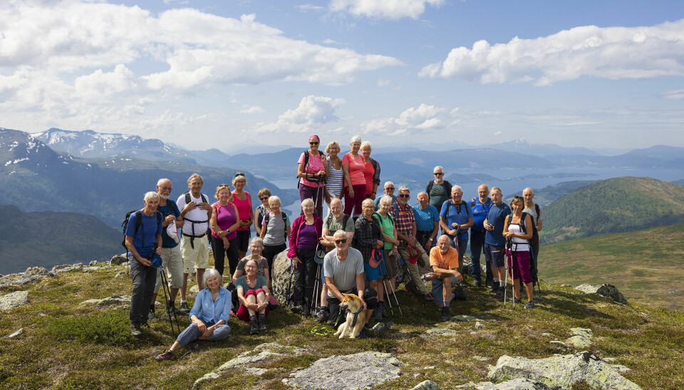 Ei gruppe med nesten 30 turkledde pensjonister oppstilt ved en varde på en fjelltopp. Fjord og fjell i bakgrunnen.