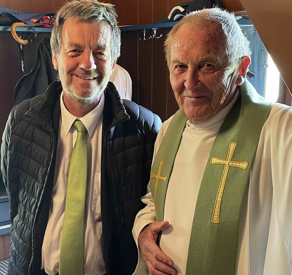 To menn, en kledd i boblejakke og grønt slips, han andre er prest og kledd i hvit skjortel med grønt bånd med kors rundt nakken