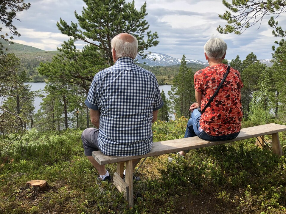 En mann og ei dame sitter på en benk med ryggen til. De har ustikt mot et vann, skog og fjell.