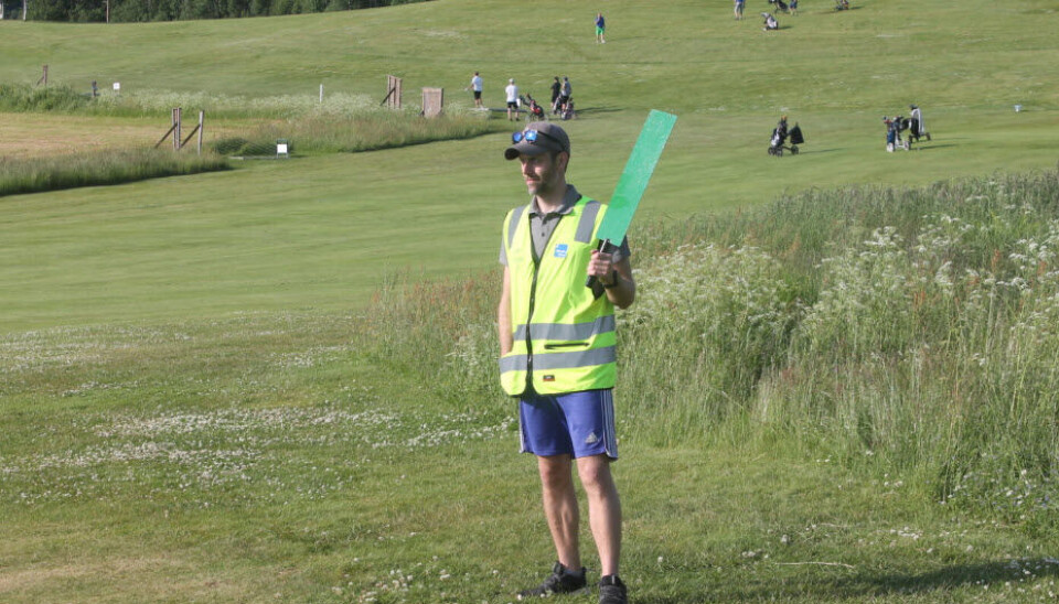 En mann i refleksvest holder opp et grønt skilt på golfbanen