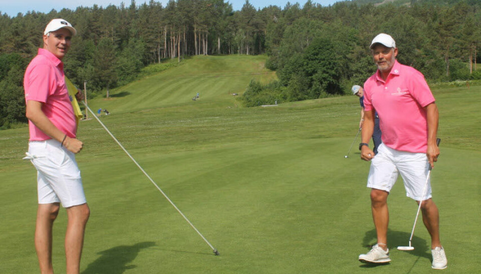 To menn kledd i rosa og hvitt står ved et golfhull og smiler mot kameraet