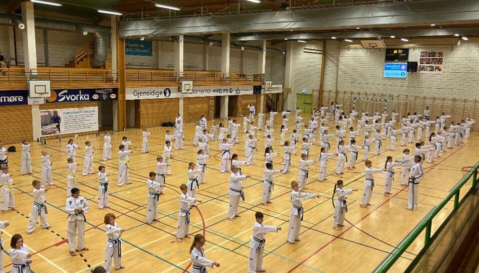 Inne i en idrettshall som er fyllt med barn som står på rekke og rad med begge føttene på gulvet og høyre hånd frem.