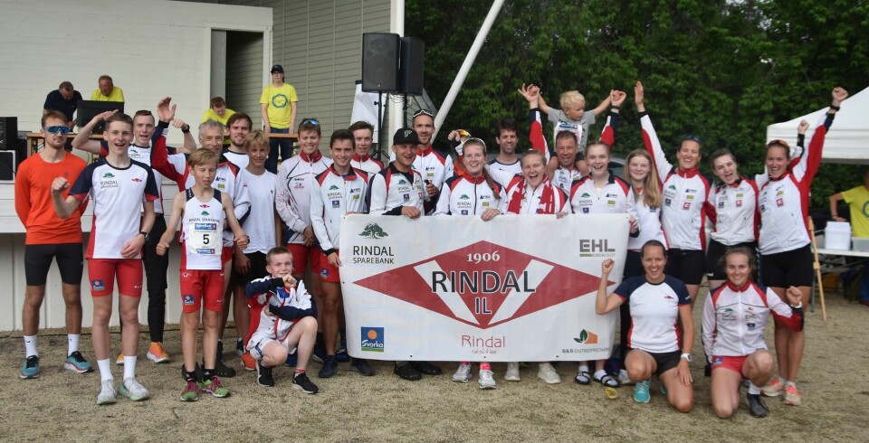 Mange menn og kvinner kledd i Rindal IL sine treningsklær. De er oppstilt og jubler og holder et flagg med idrettslagets logo foran seg.