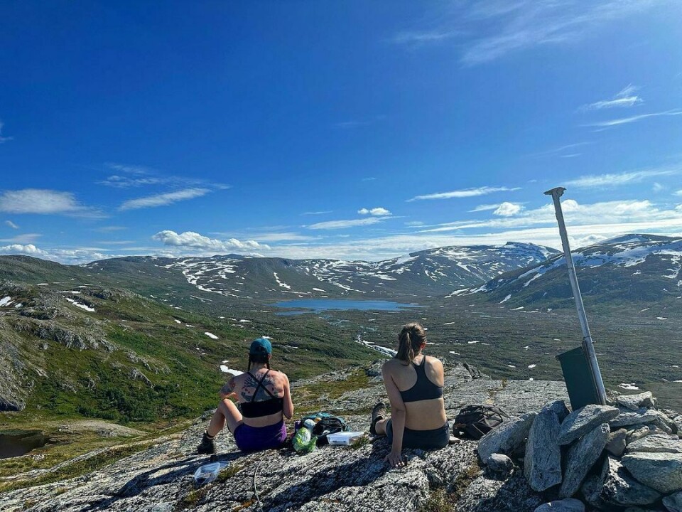 To kvinner sitter på toppen av et fjell og ser på utsikten med blå himmel og grønne fjell med litt snø