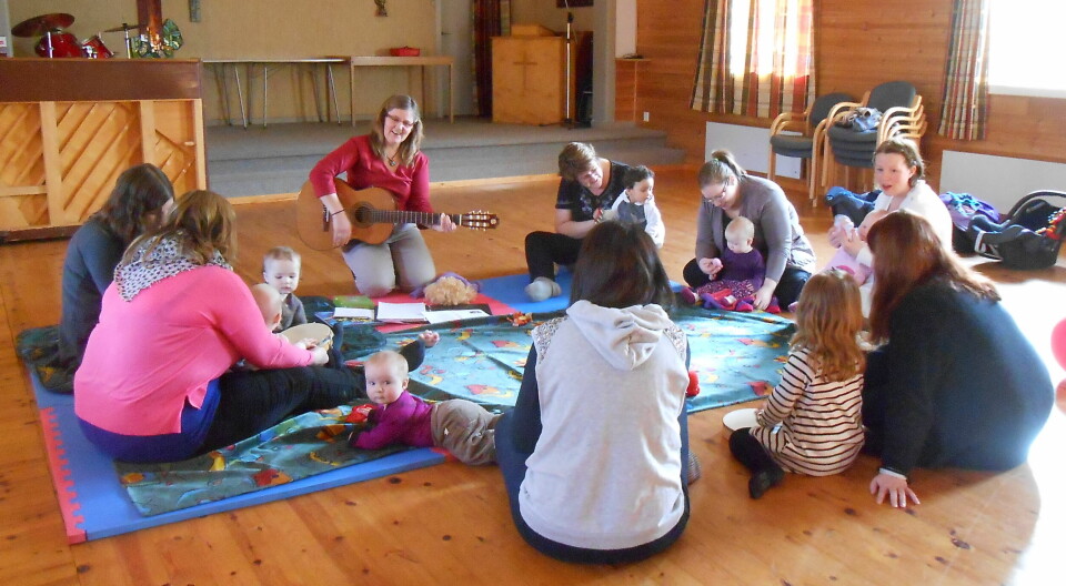 Sju mødre med små barn som sitter i ring på et teppe på gulvet, og ei dame som spiller gitar.