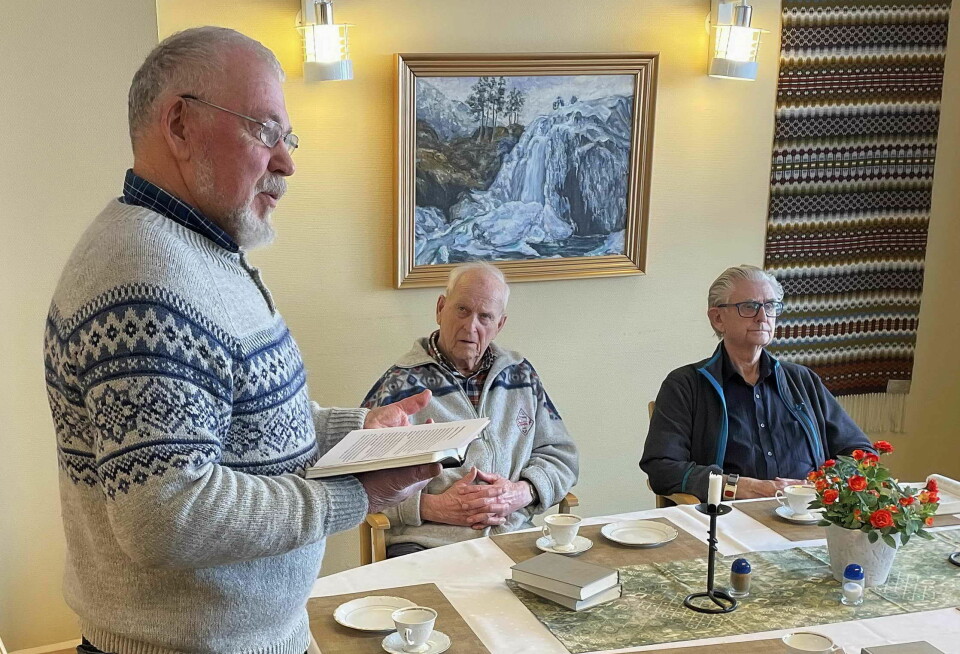 En mann i ullgenser holder en bok og snakker foran to menn som sitter ved et bord