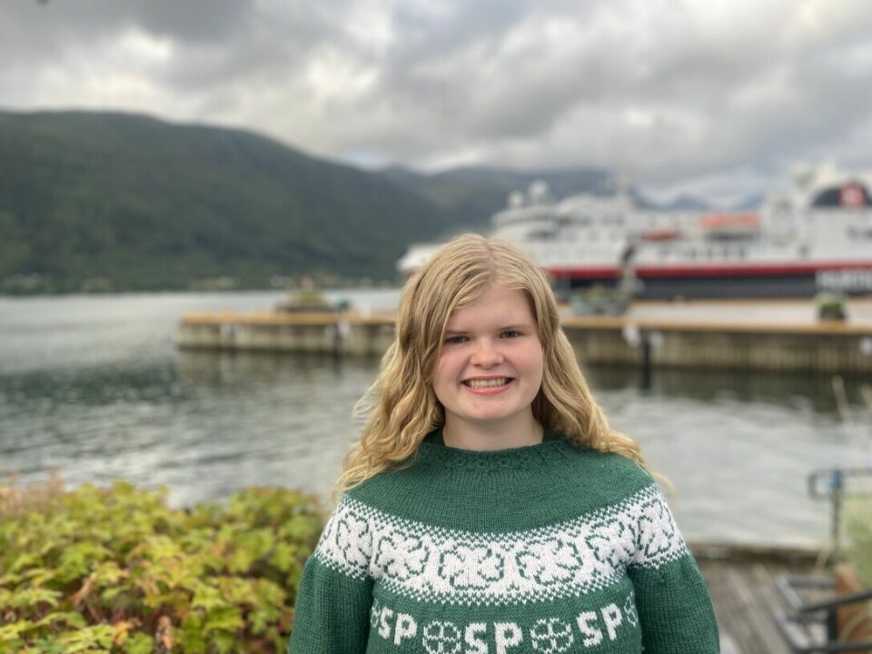 En ung blond dame står foran sjøen med en grønn strikket genser med senterpartiet sin logo