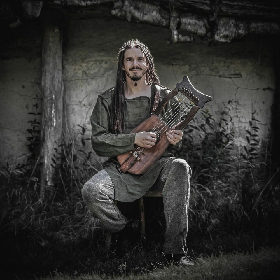 En mann med langt flettet hår og vikingklær. Han sitter på ekrekk og holder et gammeldags strengeinstrument.