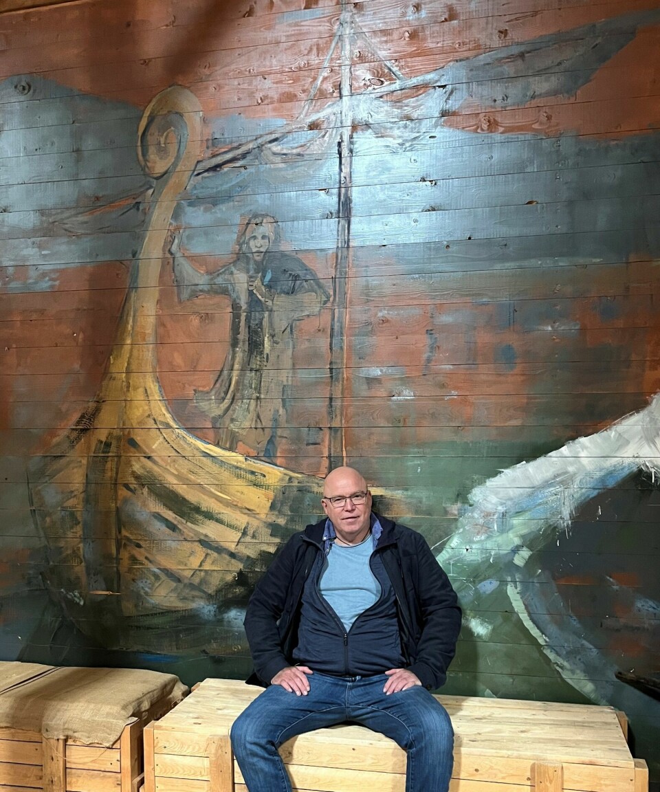 En mann som sitter på en trekasse foran en innendørs vegg der det er malt et vikingskip på veggen.