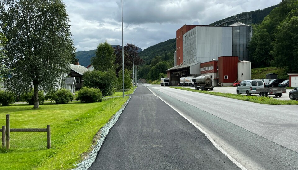 En gang- og sykkelveg med nylagt asfalt langs en asfaltert bilveg, med bolighus på ei side og et stort industribygg på den andre sida.