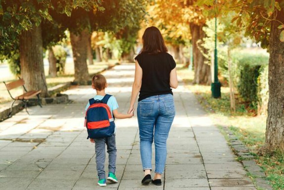 En mor holder hånden til sønnen sin som har på seg en skolesekk mens de går langs fortauet