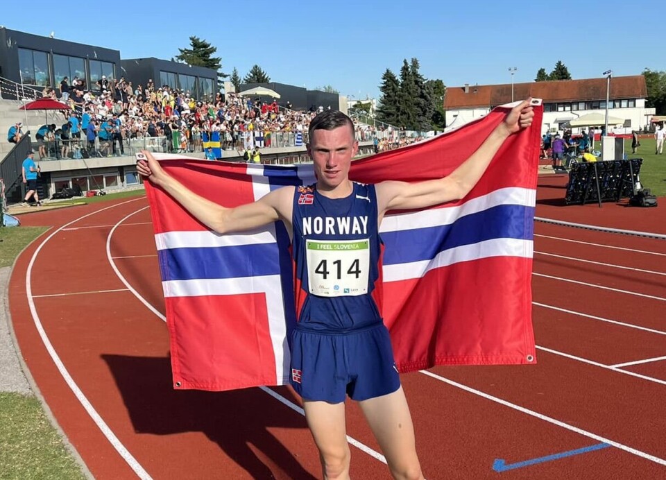 En ung mann i løperdrakt på en friidrettsstadion, med et norsk flagg bak ryggen.