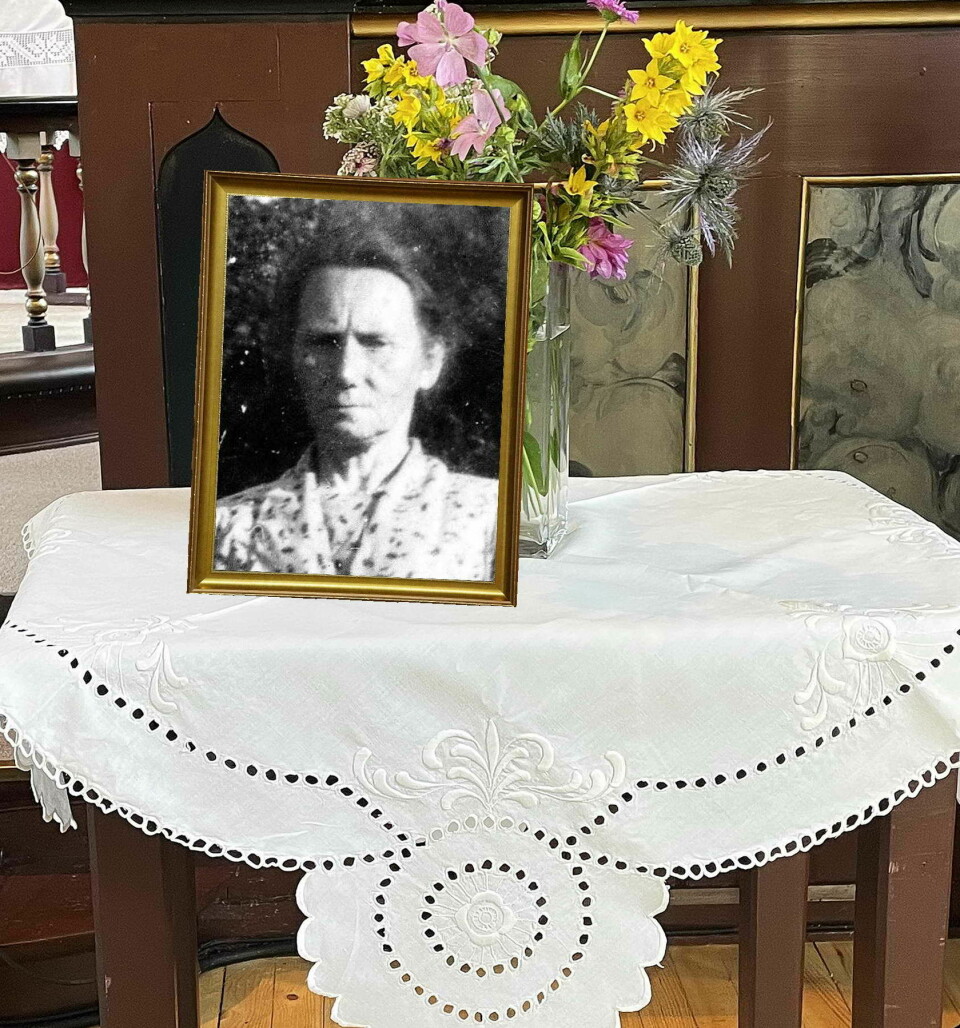 Et bord med en hvit brodert duk. På duken står en vase med blomster og et sort hvitt bilde av en eldre dame