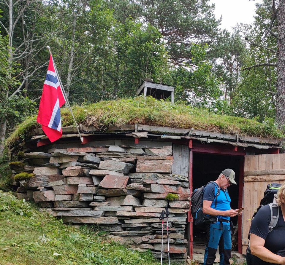 En mann står utenfor et steinhus med torvtak med et norskeflagg som henger på veggen