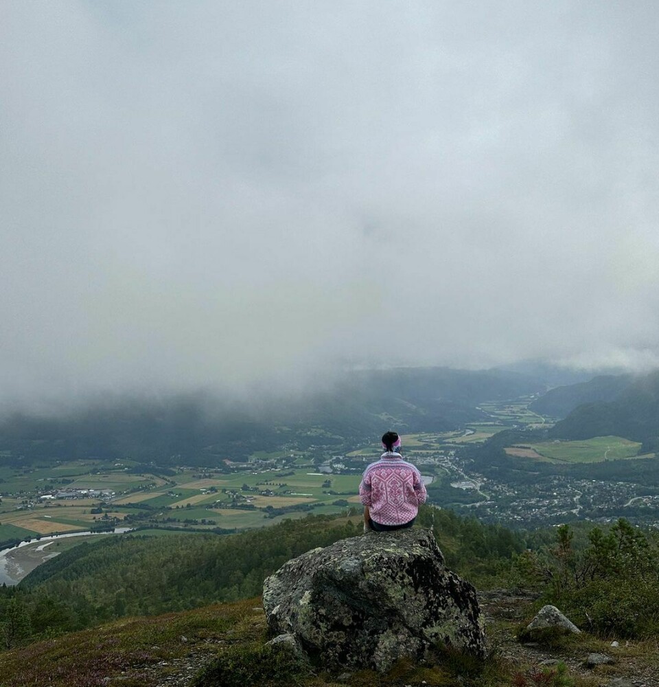 En kvinne med ullgenser sitter på en stein og ser utover utsikten ned mot bygda fra et fjell