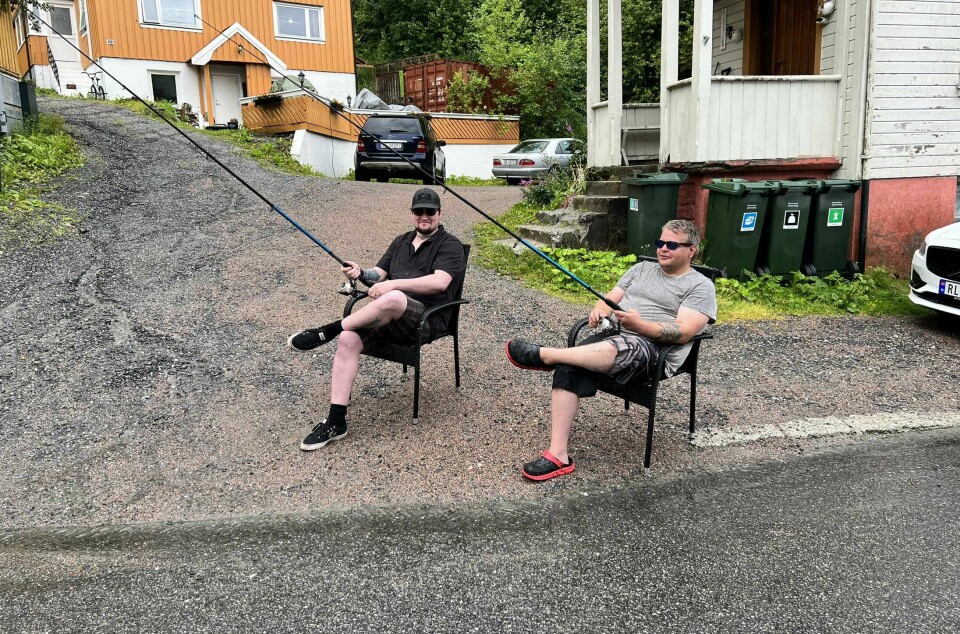 To menn sitter i en oppkjørsel i shorts og t-skjorte med hver sin fiskestang. Det har regnet mye og veien er derfor full av vann, fiskestengene ute mote veien.