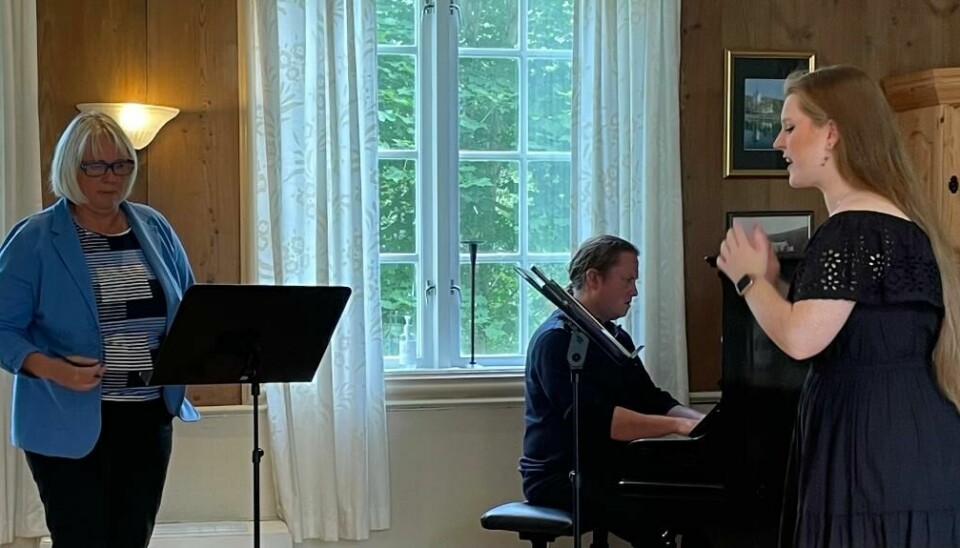 En dame synger, en mann spiller piano og en dame ser på et stativ.
