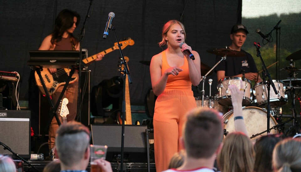 En ung dame i oransje klær synger på en scene og har et band bak seg