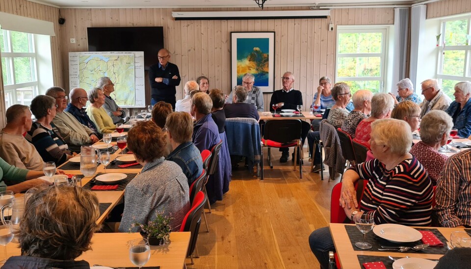 Mange personer som sitter ved langbord med tallerkner foran seg. De hører på en som står og prater.