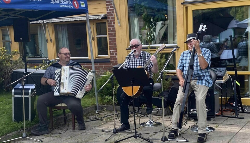 3 menn spiller musikk utendørs. Han til venstre spiller trekkspill, han i midten spiller gitar og synger og han til høyre spiller på et stort strengeinstrument.