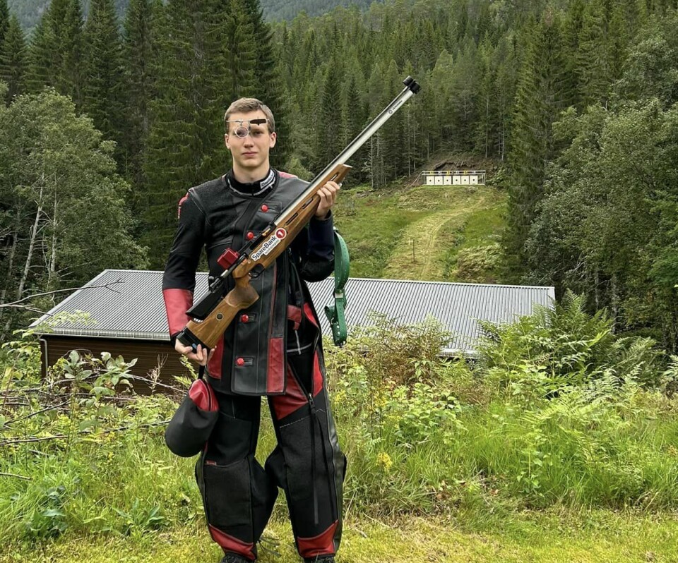 En ung mann i fullt skytterutstyr med gevær, med en utendørs skyttebane i bakgrunnen.