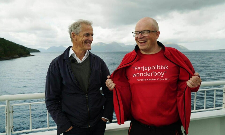 To smilende menn ved siden av hverandre med et rekkverk og sjøen i bakgrunnen. Den ene mannen er statsminister Jonas Gahr Støre. Den andre mannen er kledd i rødt. Han åpner jakken og viser t-skjorta si der det stå 'Ferjepolitisk wonderboy'.
