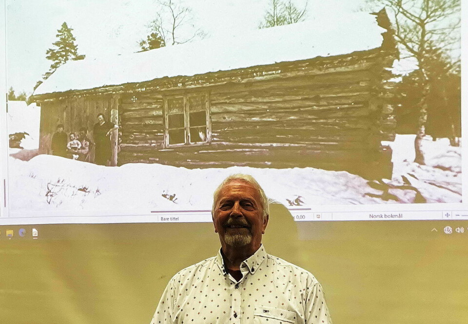 En mann med et bilde på storskjerm i bakgrunnen. Bildet viser ei gammel tømmerløe med et vindu.