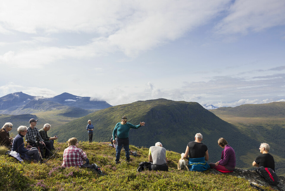 En mann står og gestikuleerer foran ei gruppe andre turkledde folk, som har satt seg ned for en rast på fjellet.