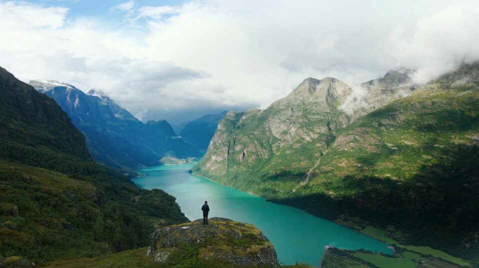 En persom som står på en fjelltopp med flere fjell rundt, og en fjord nedenfor.