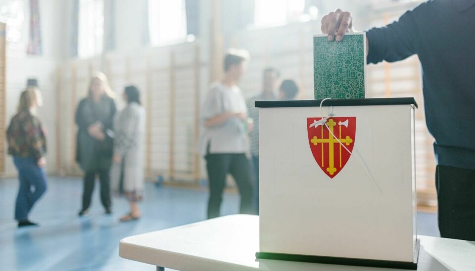 En person som putter en stemmeseddel opp i en valgurne merket med den norske kirkes våpenskjold.