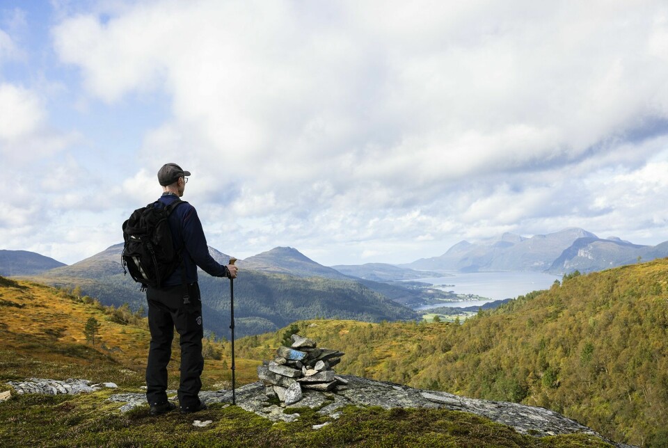 Mann med stokk står ved en liten varde og ser utover landskapet med fjell