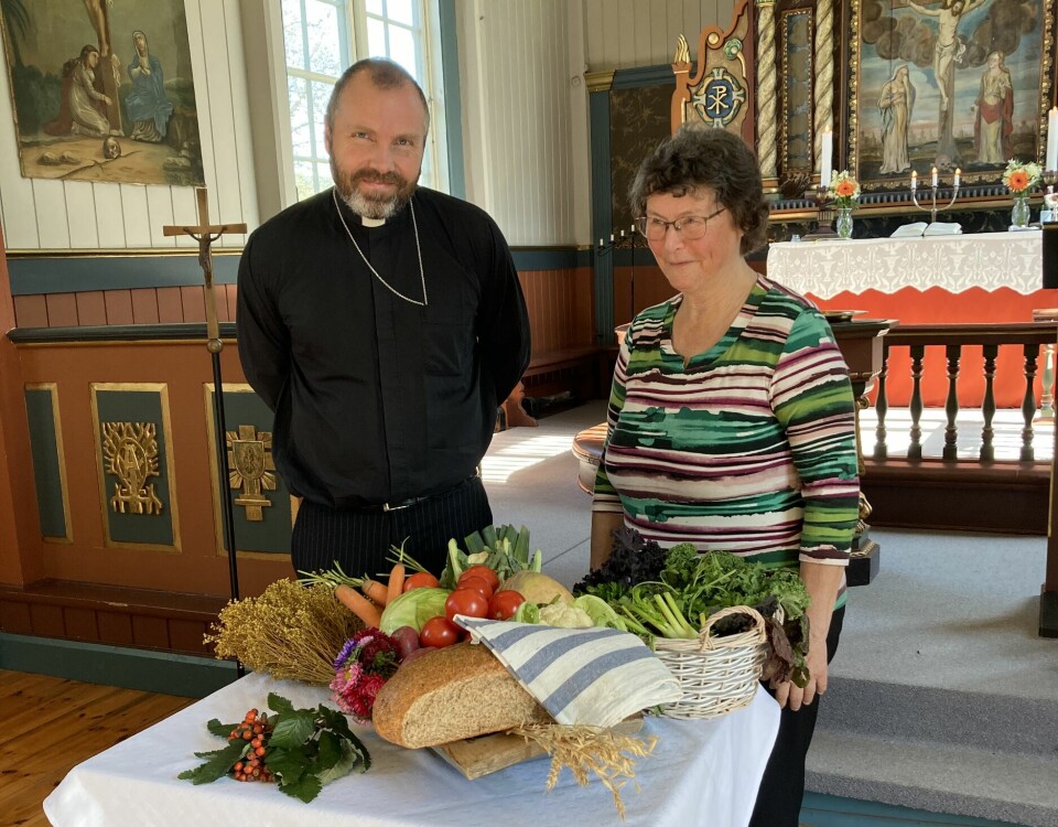 En mann i presetdrakt og ei dame som står ved et bord med brød og grønnsaker inne i ei kirke.