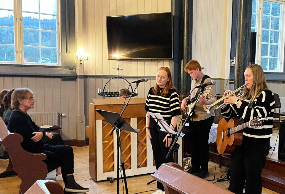 En ung gutt og to unge jenter synger og spiller gitar, bass og trompet i ei kirke.