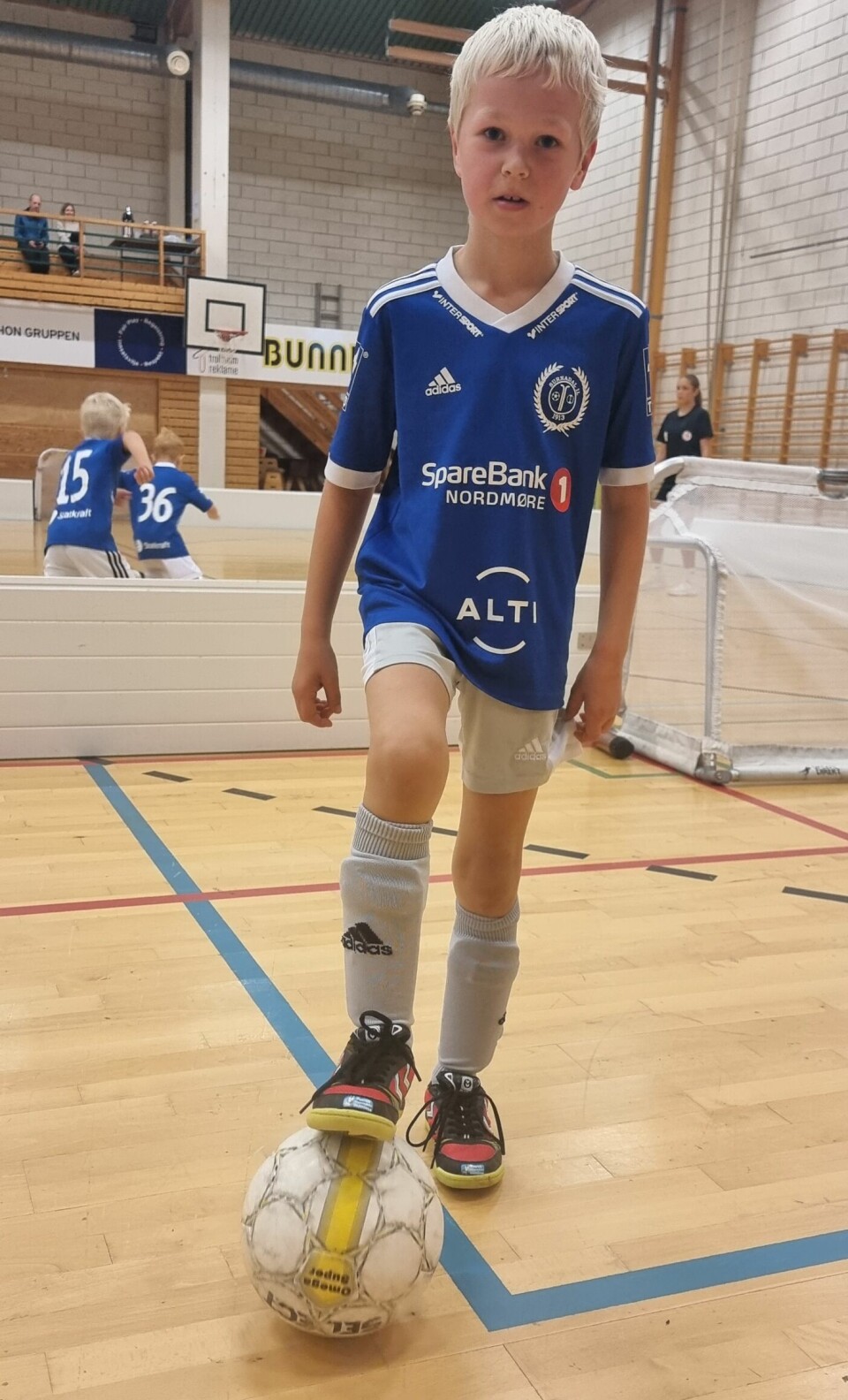 En gutt står med en foten på en fotball og har på seg en blå fotballtrøye og ser mot kamera