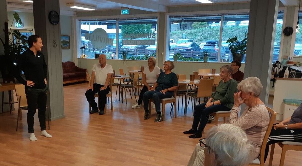 En instruktør i treningsklær står og snakker til ei gruppe pensjonister, som sitter i ring foran han på hver sin stol.
