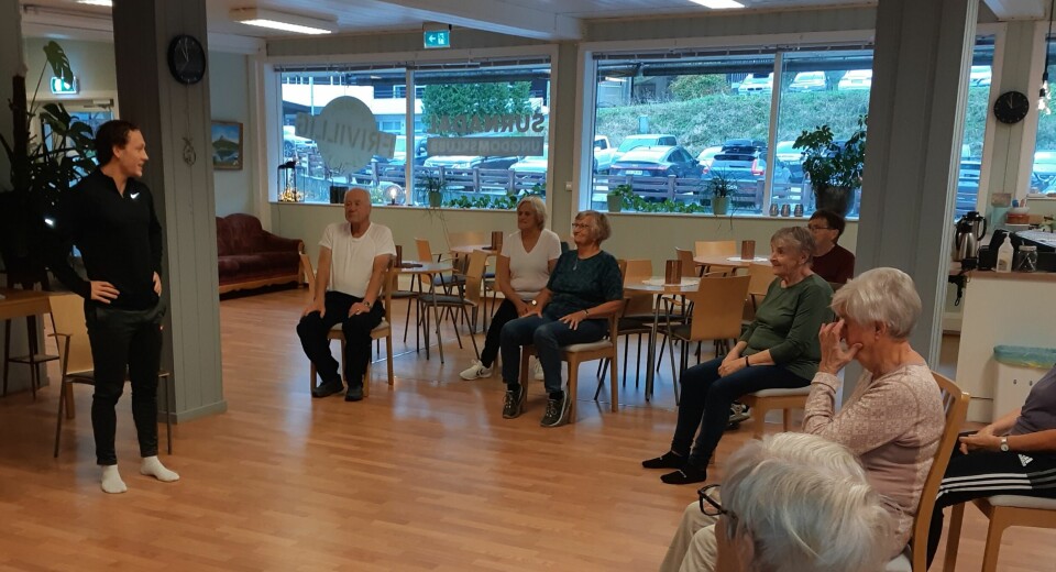 En instruktør i treningsklær står og snakker til ei gruppe pensjonister, som sitter i ring foran han på hver sin stol.
