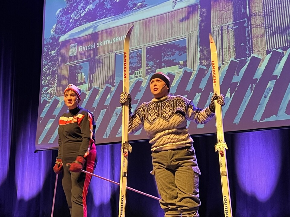 To kvinner kledd i ullgenser og lue holder ski og staver og står på en scene