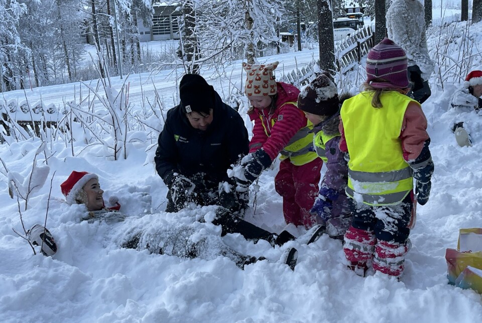 En ungdom utkledd som nisse halvveis begravd i snø. Små barn rundt han.